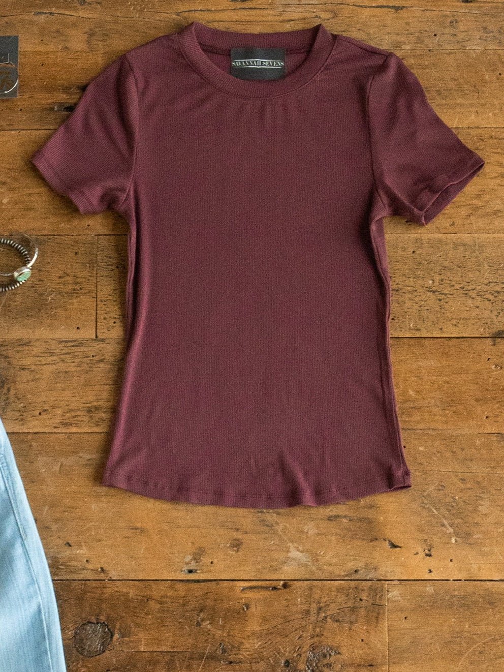 The Hadley Rosewood Ribbed Basic Tee by Savannah Sevens western life{&}style is displayed flat on a wooden surface, alongside a glimpse of a light blue garment and a bracelet.