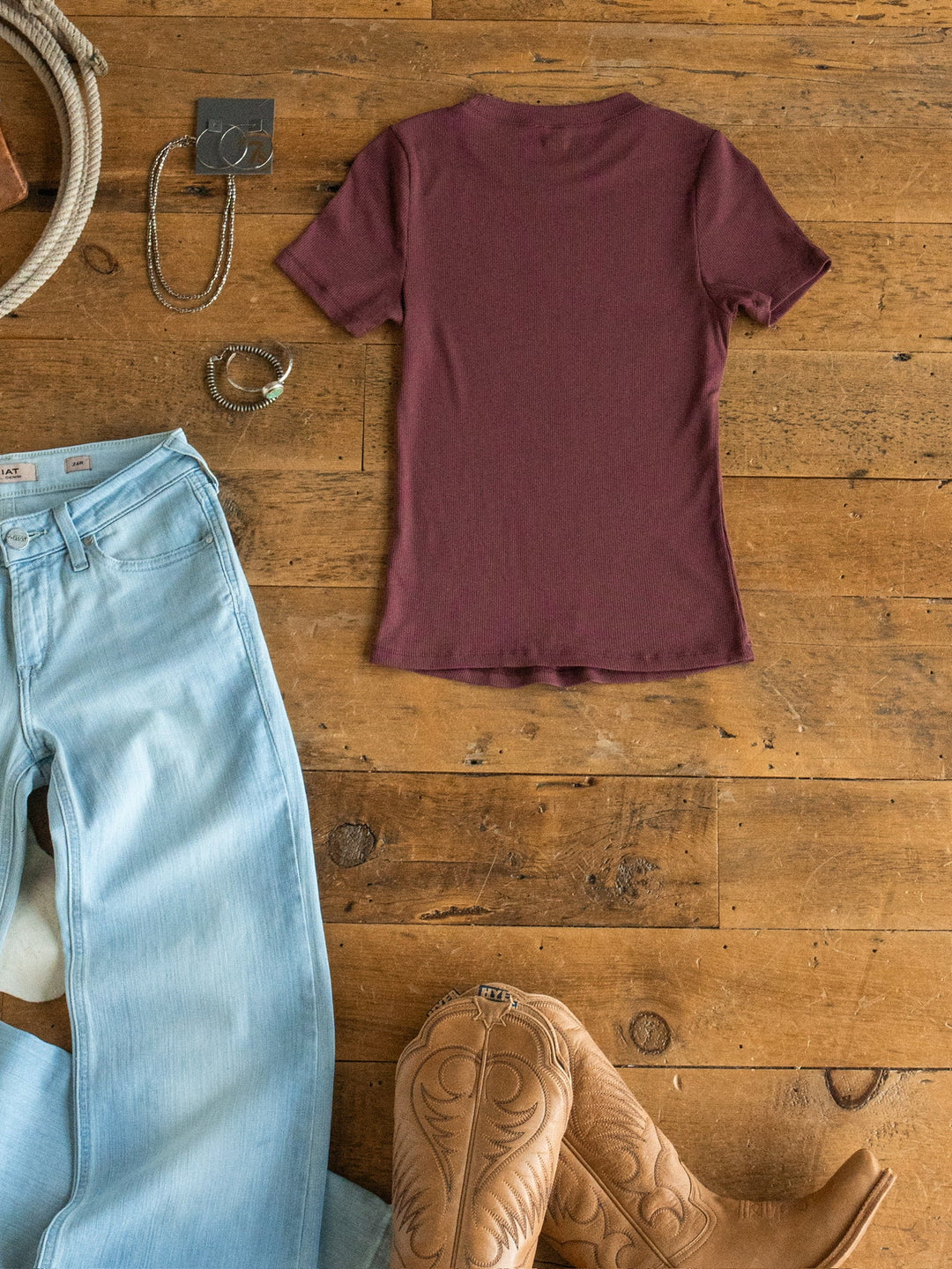 A flat lay featuring the Hadley Rosewood Ribbed Basic Tee by Savannah Sevens western life{&}style arranged with light blue jeans, tan cowboy boots, a silver belt buckle, a bracelet, and a necklace on a wooden floor.