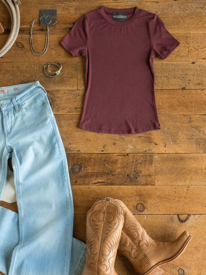 A flat lay featuring the Hadley Rosewood Ribbed Basic Tee by Savannah Sevens western life{&}style, paired with light blue jeans, tan cowboy boots, a silver bracelet, a ring, and a rope displayed on a wooden floor.