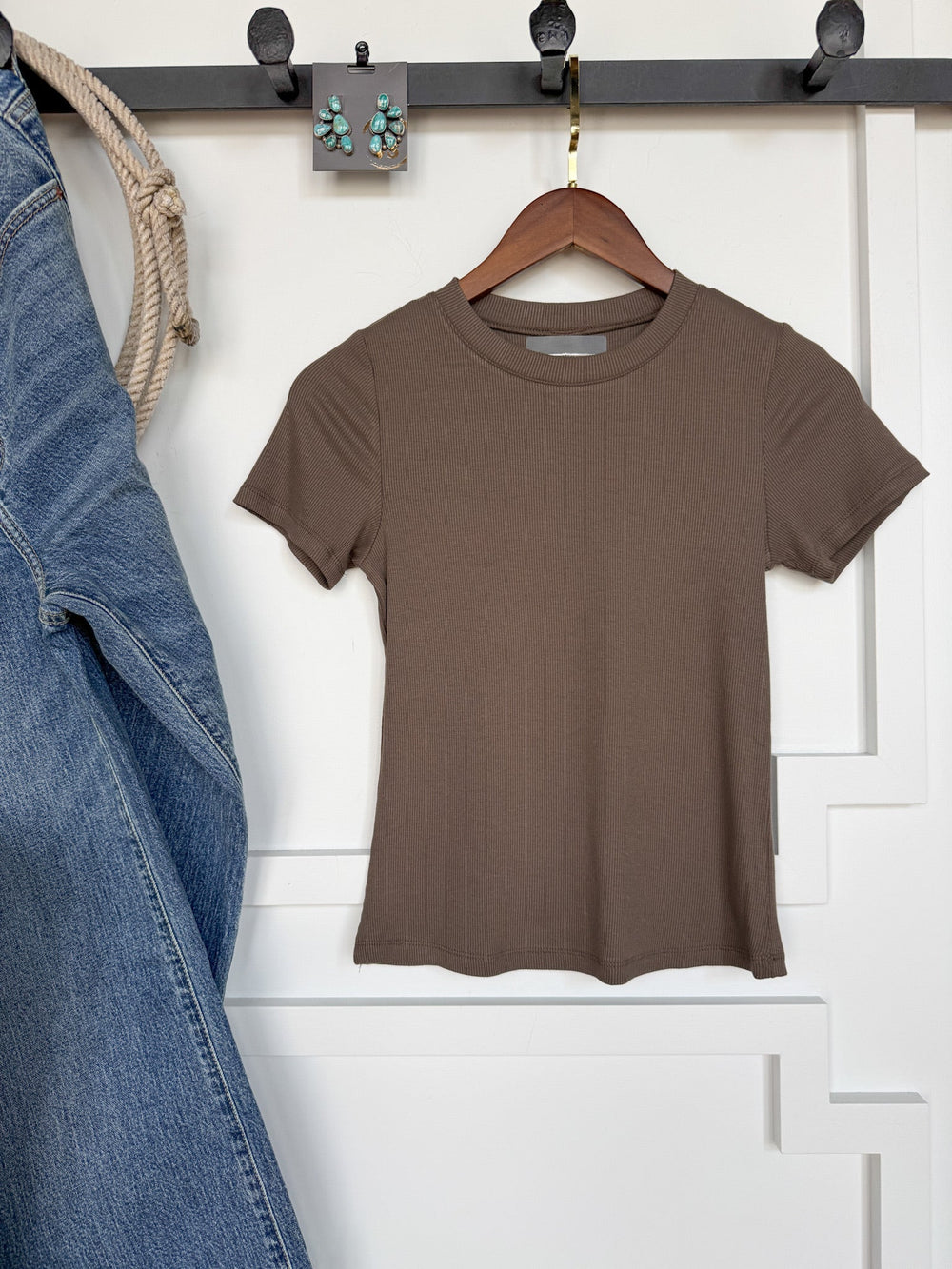 The Hadley Truffle Ribbed Basic Tee by Savannah Sevens western life{&}style hangs on a wooden hanger against a white wall, displayed next to a denim jacket and statement earrings on a black hook rack.