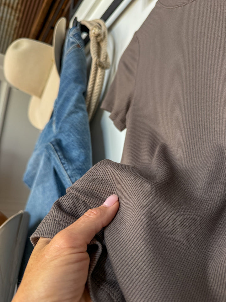 A hand holding the Hadley Truffle Ribbed Basic Tee from Savannah Sevens western life{&}style, with a pair of blue jeans draped over a chair and a beige hat hanging in the background.