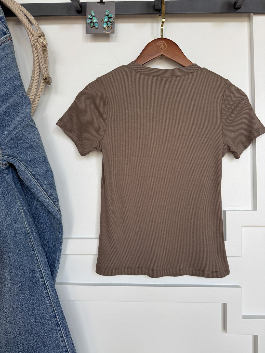 The Hadley Truffle Ribbed Basic Tee by Savannah Sevens western life{&}style hangs on a wooden hanger against a white wall. Blue jeans are displayed to the left, with a rope looped above. Green and silver earrings are arranged on the wall above the shirt.