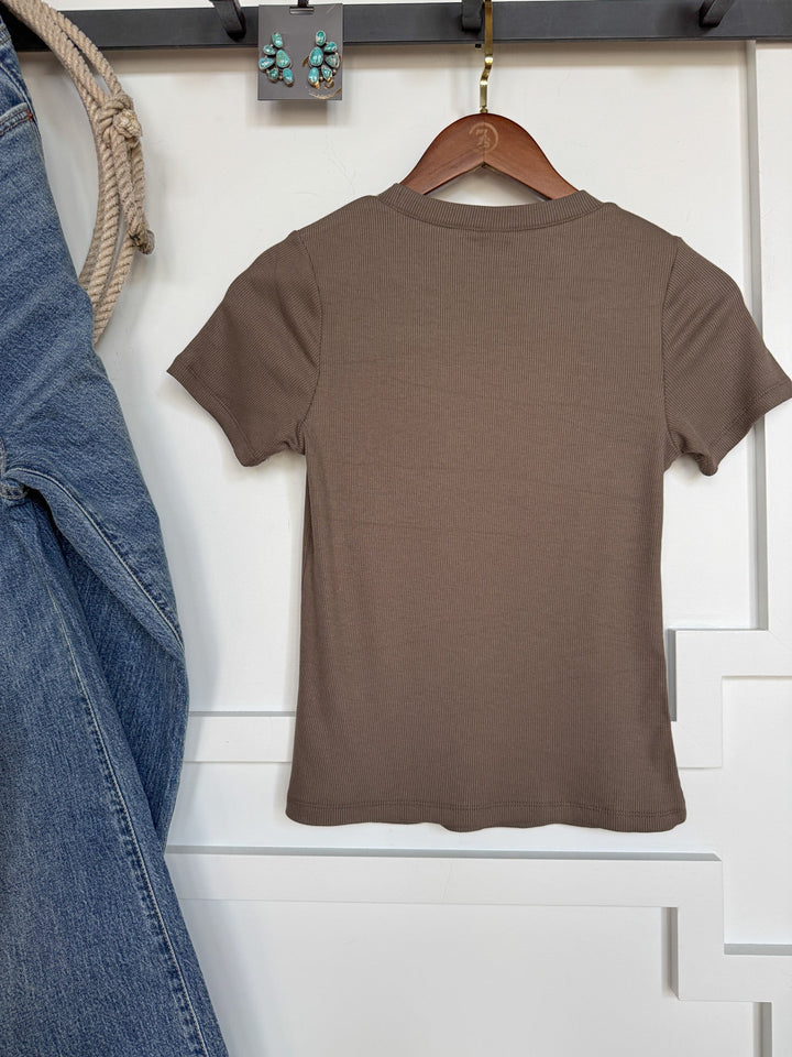 The Hadley Truffle Ribbed Basic Tee by Savannah Sevens western life{&}style hangs on a wooden hanger against a white wall. Blue jeans are displayed to the left, with a rope looped above. Green and silver earrings are arranged on the wall above the shirt.
