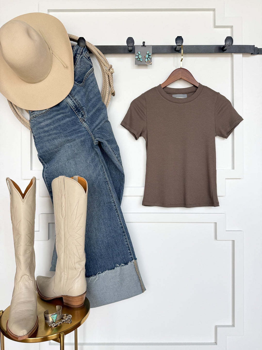 The Hadley Truffle Ribbed Basic Tee by Savannah Sevens western life{&}style is styled with a tan cowboy hat, turquoise earrings, and denim jeans hanging on a wall rack. Cream cowboy boots and jewelry are displayed on a small gold table below, creating a stylish Western-inspired outfit arrangement in young contemporary sizing.