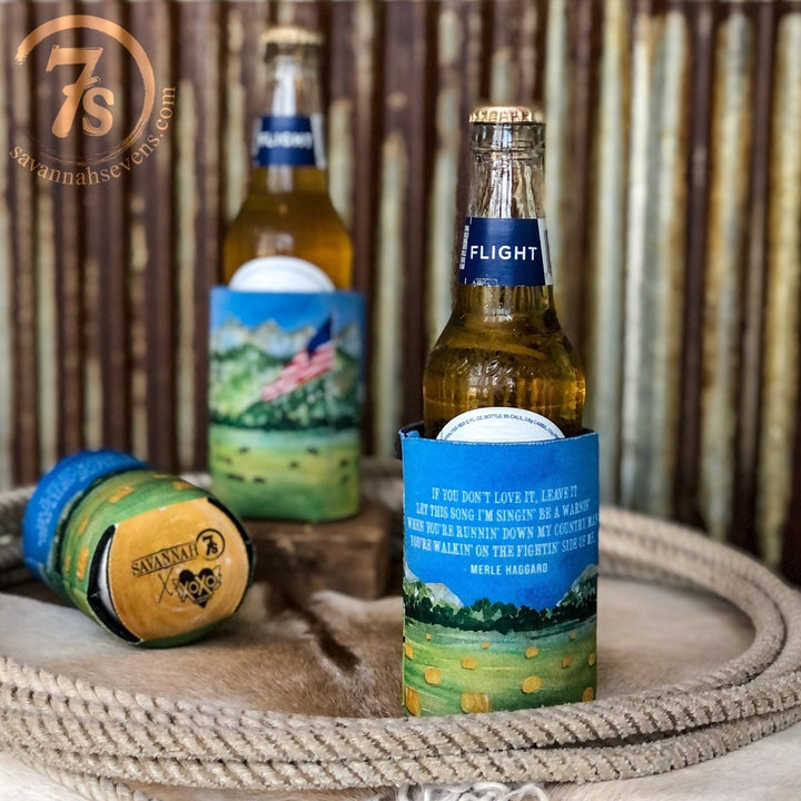 A bottle of beer in a countryside-themed Haggard Koozie by Savannah Sevens western life{&}style rests on a rope-wrapped surface, with another bottle and branded koozie in the background. The Savannah 7s logo appears in the top left corner.