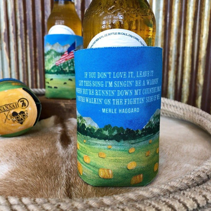 A beer bottle in the Haggard Koozie by Savannah Sevens western life{&}style, with a scenic landscape and Merle Haggard quote. Behind it, another bottle sports an American flag koozie—perfect for capturing the Savannah 7s country vibe.