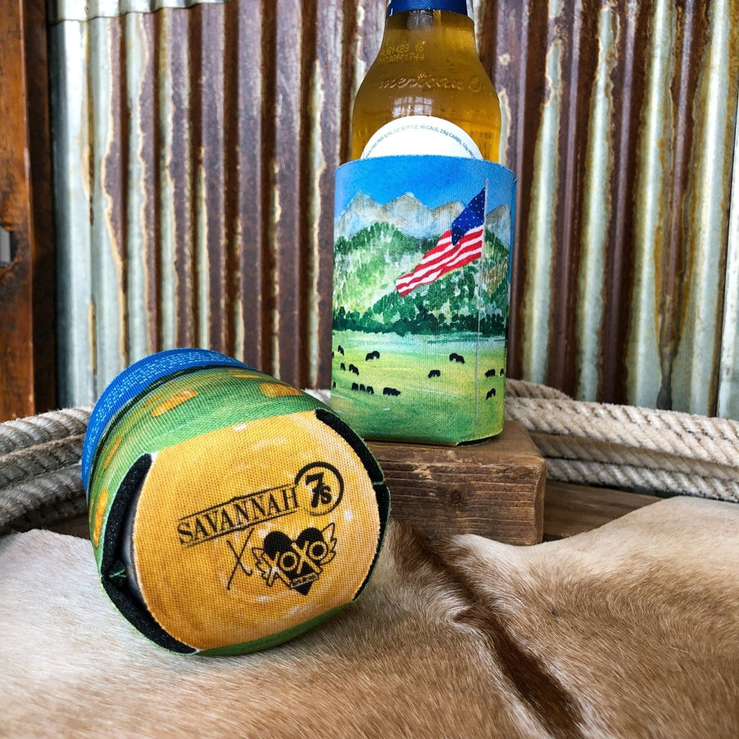 A Haggard Koozie by Savannah Sevens western life{&}style, featuring a mountain and flag design, stands upright beside another sleeve with "SAVANNAH 73 XOXO" text. Both rest on a surface in front of corrugated metal and a rope.