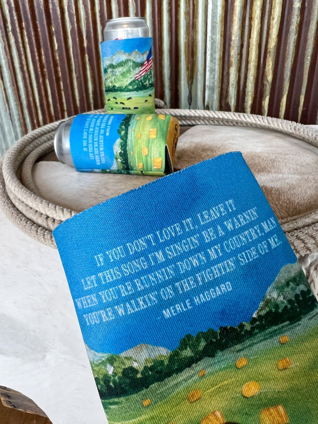 A close-up of the Haggard Koozie by Savannah Sevens western life{&}style, showing country landscape art and Merle Haggard lyrics, in front of similar cans, all set on a coiled rope and cowhide with corrugated metal behind.