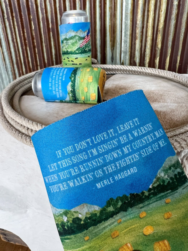A close-up of the Haggard Koozie by Savannah Sevens western life{&}style, showing country landscape art and Merle Haggard lyrics, in front of similar cans, all set on a coiled rope and cowhide with corrugated metal behind.
