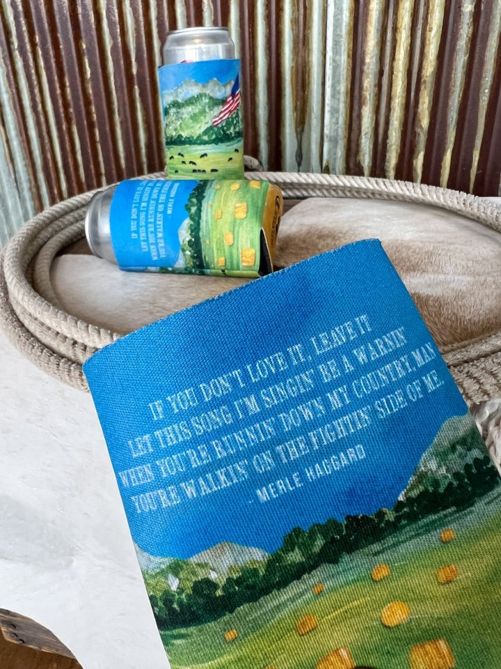 A close-up of the Haggard Koozie by Savannah Sevens western life{&}style, showing country landscape art and Merle Haggard lyrics, in front of similar cans, all set on a coiled rope and cowhide with corrugated metal behind.
