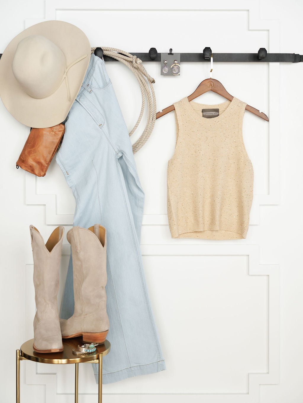 A beige wide-brim hat, light-wash jeans, Halsey Natural Woven Sleeveless Top by Savannah Sevens western life{&}style, leather purse, and rope hang on a black rack. Below, tan cowboy boots and jewelry rest on a gold table near a white wall.