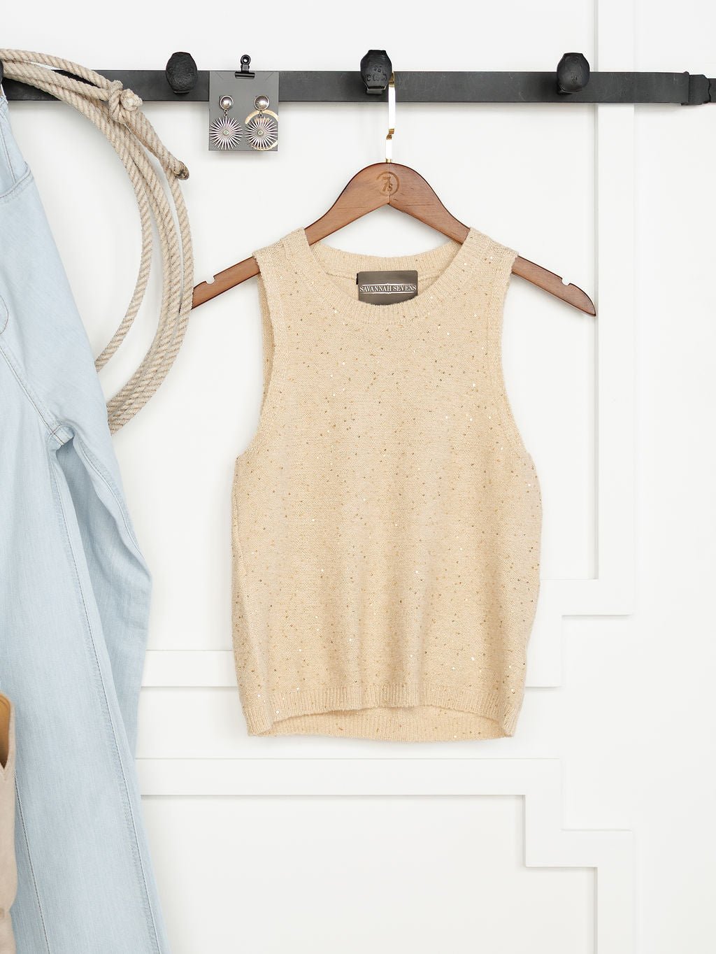 The Halsey Natural Woven Sleeveless Top by Savannah Sevens western life{&}style hangs on a wooden hanger against a white wall, with a light blue denim jacket, coiled rope, and black rack holding earrings nearby.