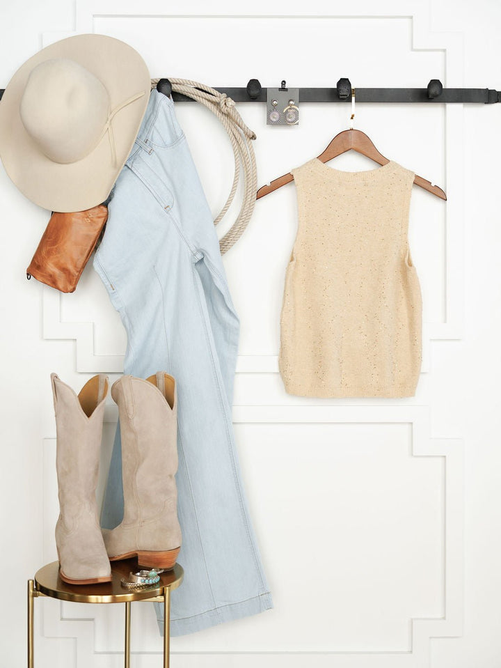 A wide-brimmed hat, brown clutch, light blue jeans, and a beige knit tank top hang on hooks above a table with suede cowboy boots and turquoise jewelry. The Halsey Natural Woven Sleeveless Top by Savannah Sevens adds natural shimmer to the white paneled wall.