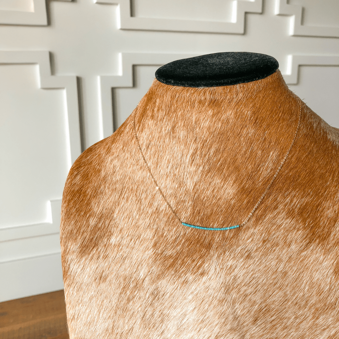 The Halsey Necklace by Savannah Sevens western life{&}style features a dainty gold filled chain with a small horizontal turquoise bar, shown on a brown and white cowhide bust against a white paneled wall.