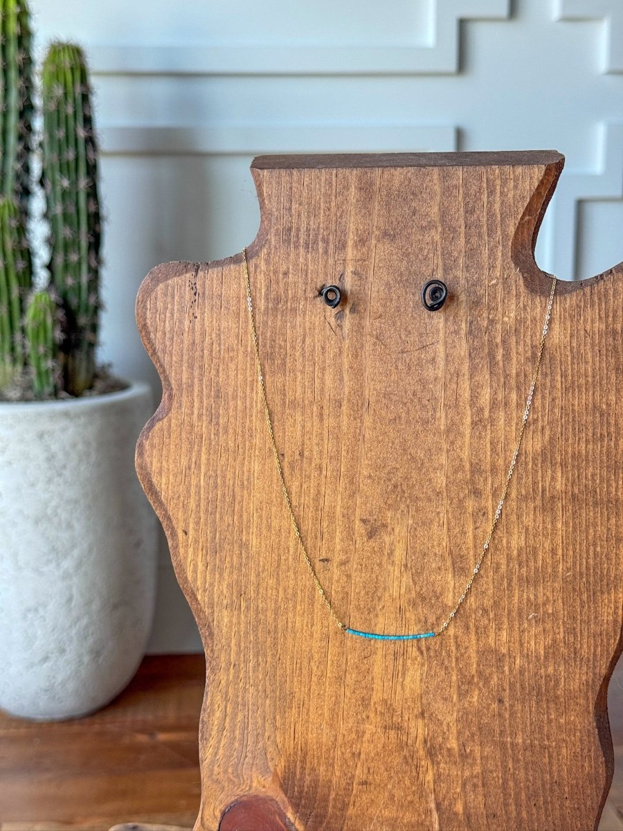 The Halsey Necklace by Savannah Sevens western life{&}style—a delicate turquoise bar with small beads on a gold filled chain—is displayed on a wooden jewelry bust, with a tall potted cactus in the background against a white paneled wall.
