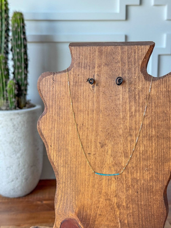The Halsey Necklace by Savannah Sevens western life{&}style—a delicate turquoise bar with small beads on a gold filled chain—is displayed on a wooden jewelry bust, with a tall potted cactus in the background against a white paneled wall.