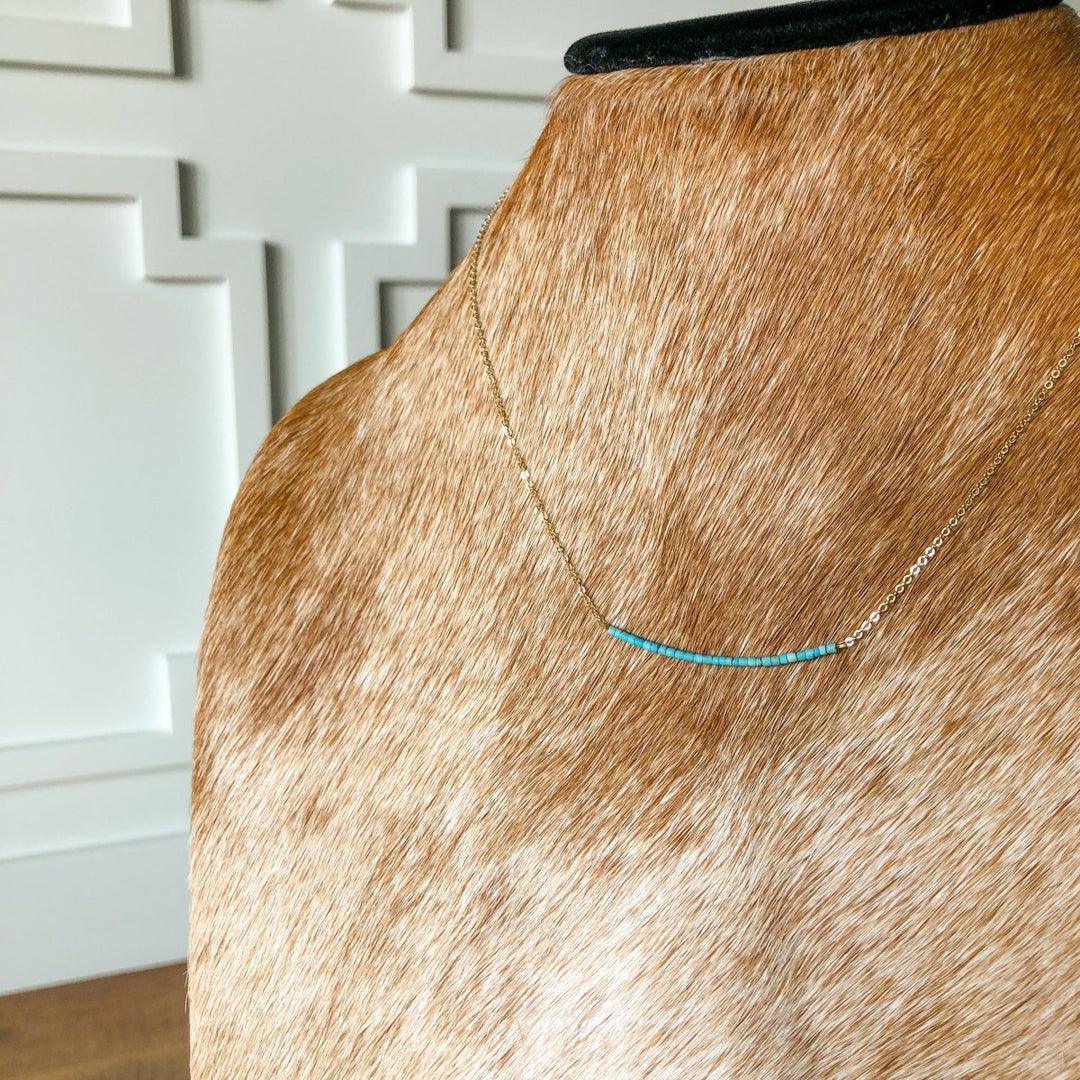 The Halsey Necklace by Savannah Sevens western life{&}style is a dainty gold filled piece with a turquoise bar and small beads, shown on a light brown cowhide bust against a white paneled wall.