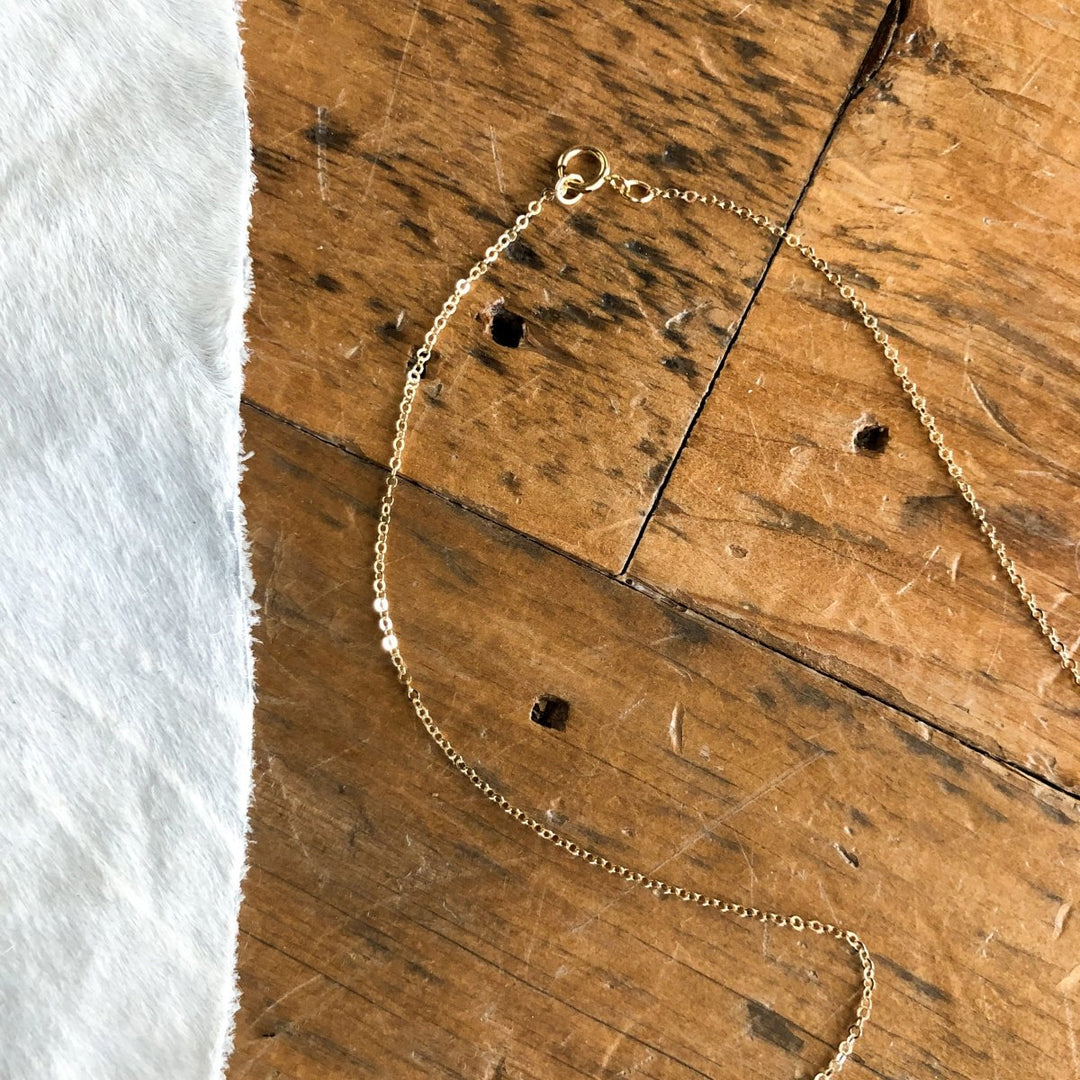 The Halsey Necklace by Savannah Sevens western life{&}style, a delicate gold-filled piece with a clasp, is displayed on a rustic wooden surface beside white cowhide.