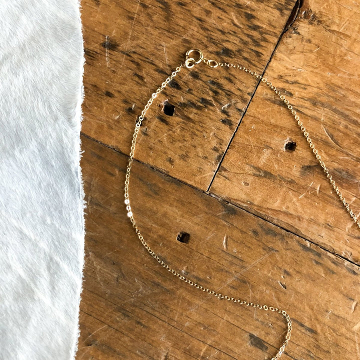 The Halsey Necklace by Savannah Sevens western life{&}style, a delicate gold-filled piece with a clasp, is displayed on a rustic wooden surface beside white cowhide.