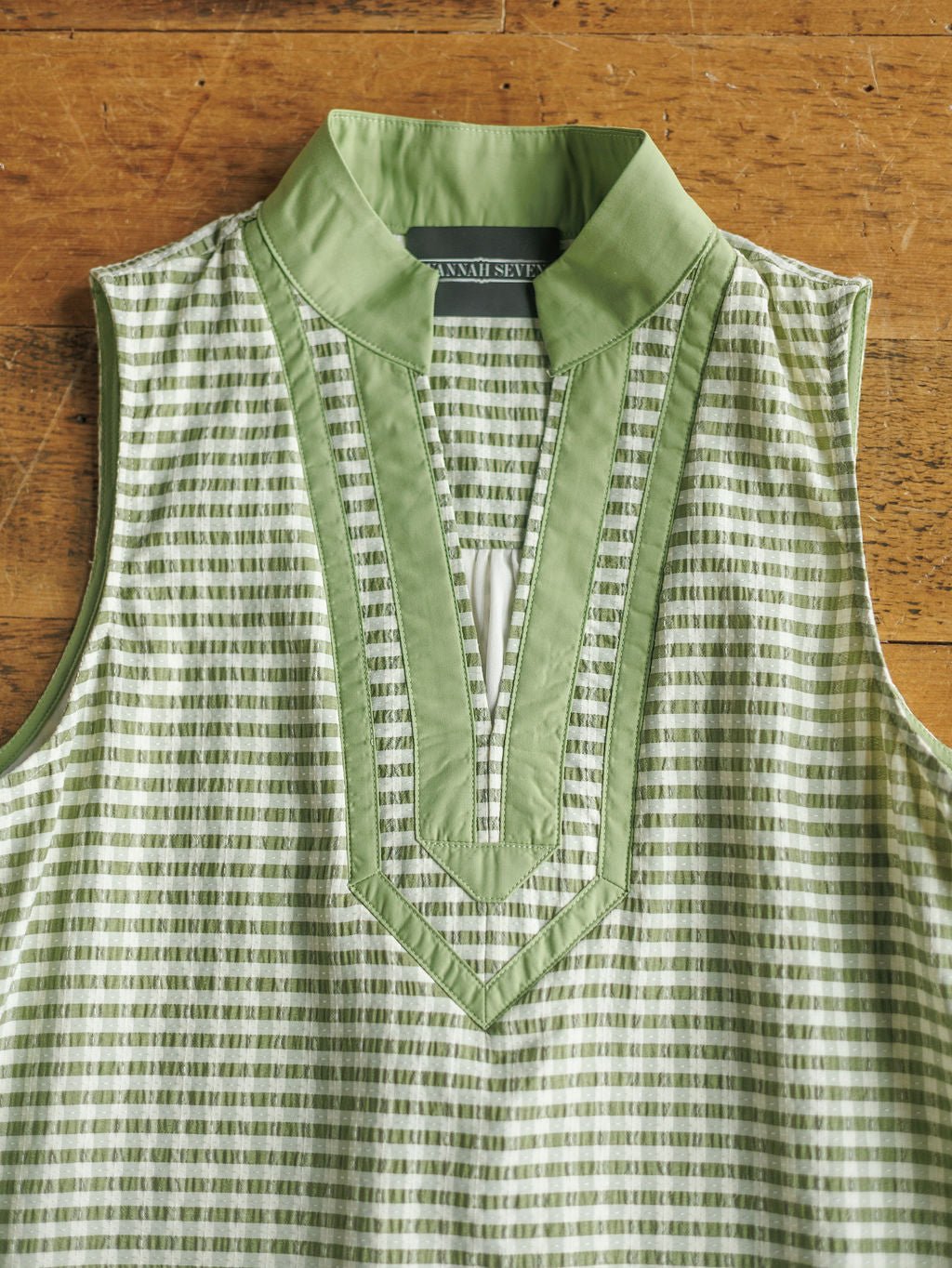 The Halstad Sage Gingham Dress by Savannah Sevens western life{&}style, featuring a sleeveless green and white checkered design with a V-neck and green collar, is displayed flat on a wooden surface.