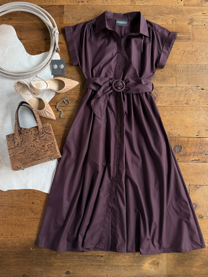 The Hanley Hills Fig Button Up Dress by Savannah Sevens western life{&}style, a short-sleeve purple cotton blend dress with a belt, is laid out on wood beside beige heels, a cork handbag, two rope coils, and a pair of black earrings on a card.