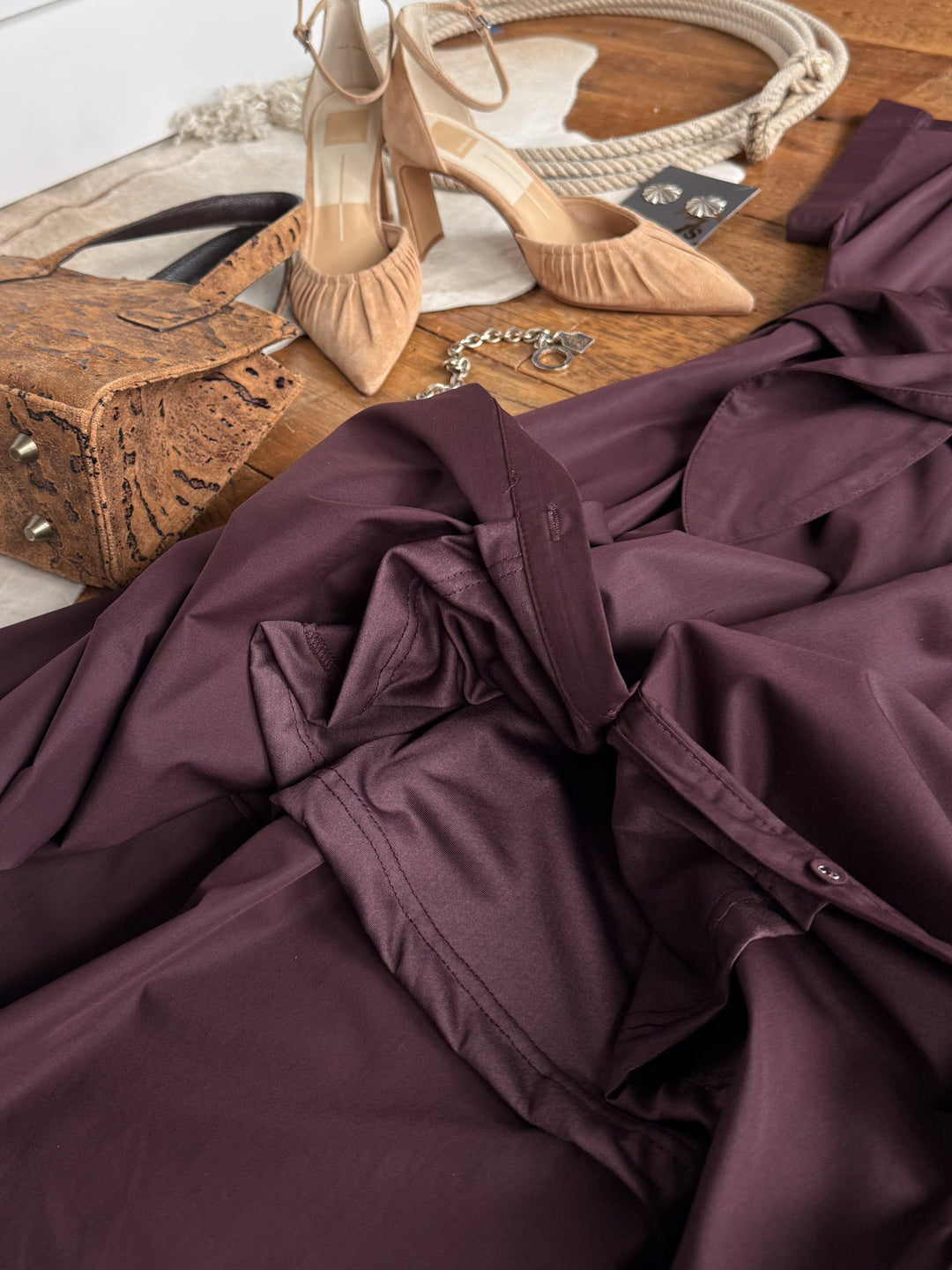 The Hanley Hills Fig Button Up Dress by Savannah Sevens western life{&}style, a cotton blend short sleeve midi dress in burgundy, is styled in a flat lay on a wooden floor alongside tan high heels, a brown textured purse, a necklace, and black earrings for an effortlessly chic look.