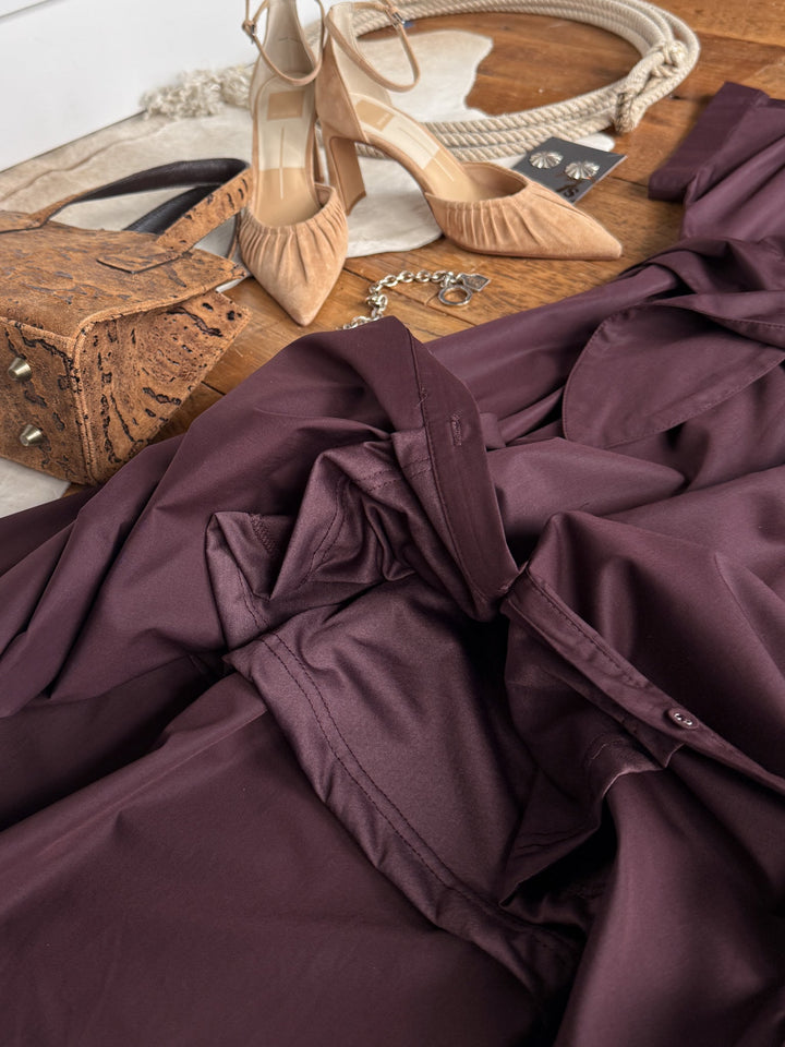 The Hanley Hills Fig Button Up Dress by Savannah Sevens western life{&}style, a cotton blend short sleeve midi dress in burgundy, is styled in a flat lay on a wooden floor alongside tan high heels, a brown textured purse, a necklace, and black earrings for an effortlessly chic look.