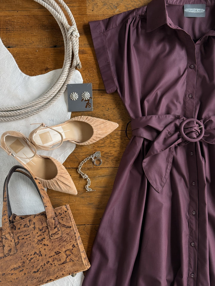 A flat lay featuring the Hanley Hills Fig Button Up Dress by Savannah Sevens western life{&}style—a short sleeve midi dress in maroon—styled with beige pointed heels, a cork handbag, silver earrings, a bracelet, and a large rope, all arranged on a cowhide rug over a wooden floor.