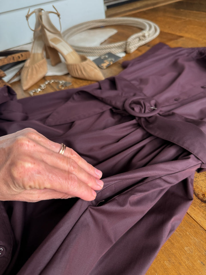 A hand with a ring touches the pocket of the Hanley Hills Fig Button Up Dress by Savannah Sevens western life{&}style, laid on a wooden surface, with beige shoes, jewelry, and accessories in the background.