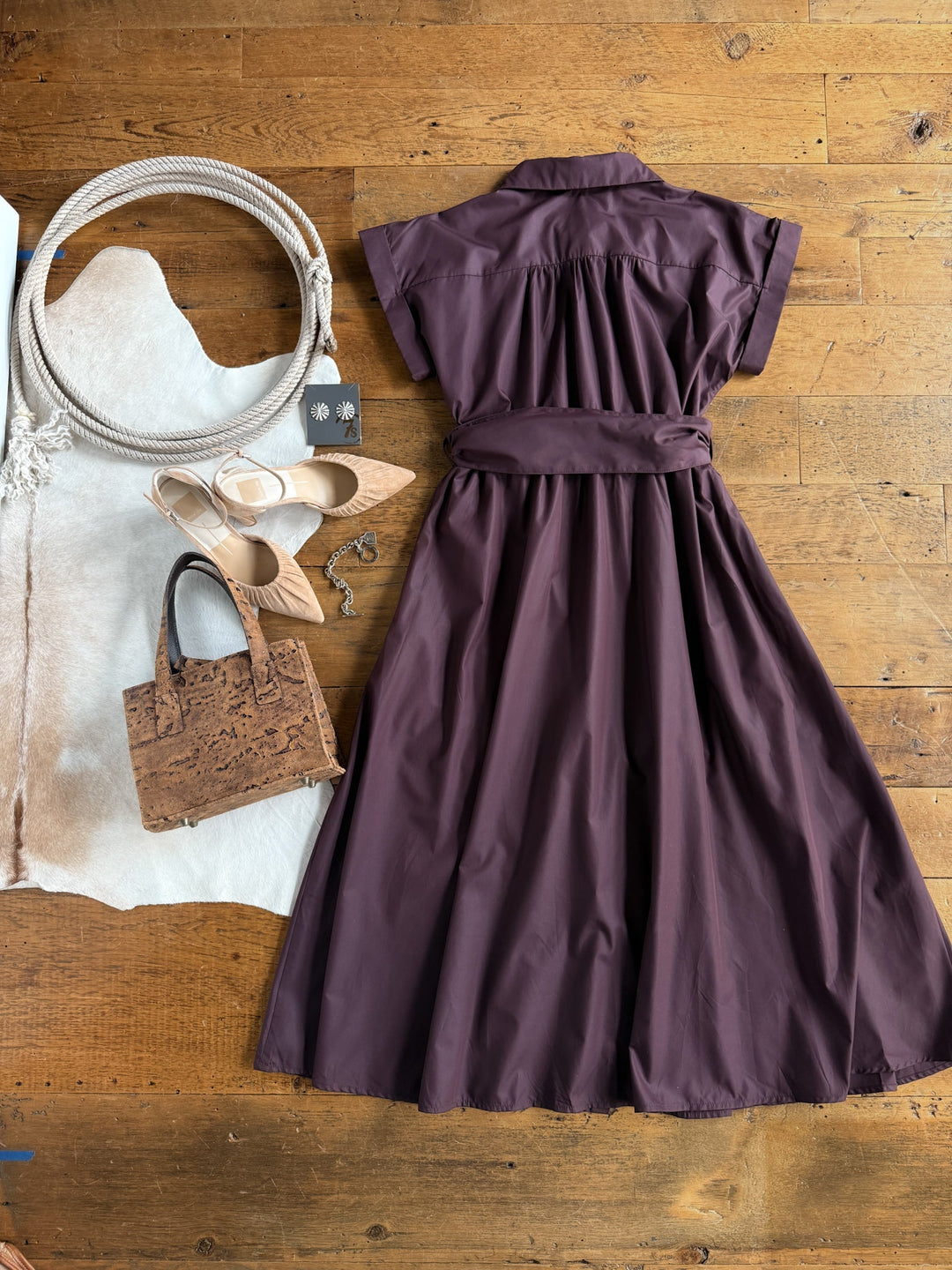 The Hanley Hills Fig Button Up Dress by Savannah Sevens western life{&}style—a purple cotton blend short-sleeve midi dress with a belt—is laid out on a wooden floor, next to a brown handbag, cream sandals, a white rope, and part of a white cowhide rug.