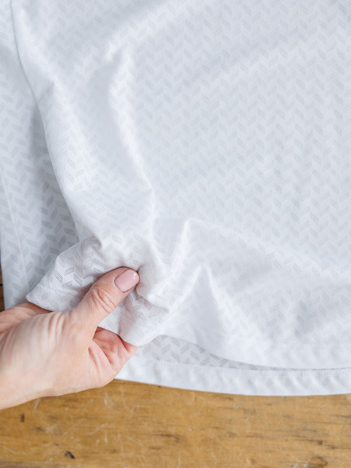 A hand with light pink nail polish pinches the edge of the Hanover Ariat White Golf Tank by Savannah Sevens western life{&}style, showing its breathable white fabric with a subtle chevron pattern on a wooden surface.