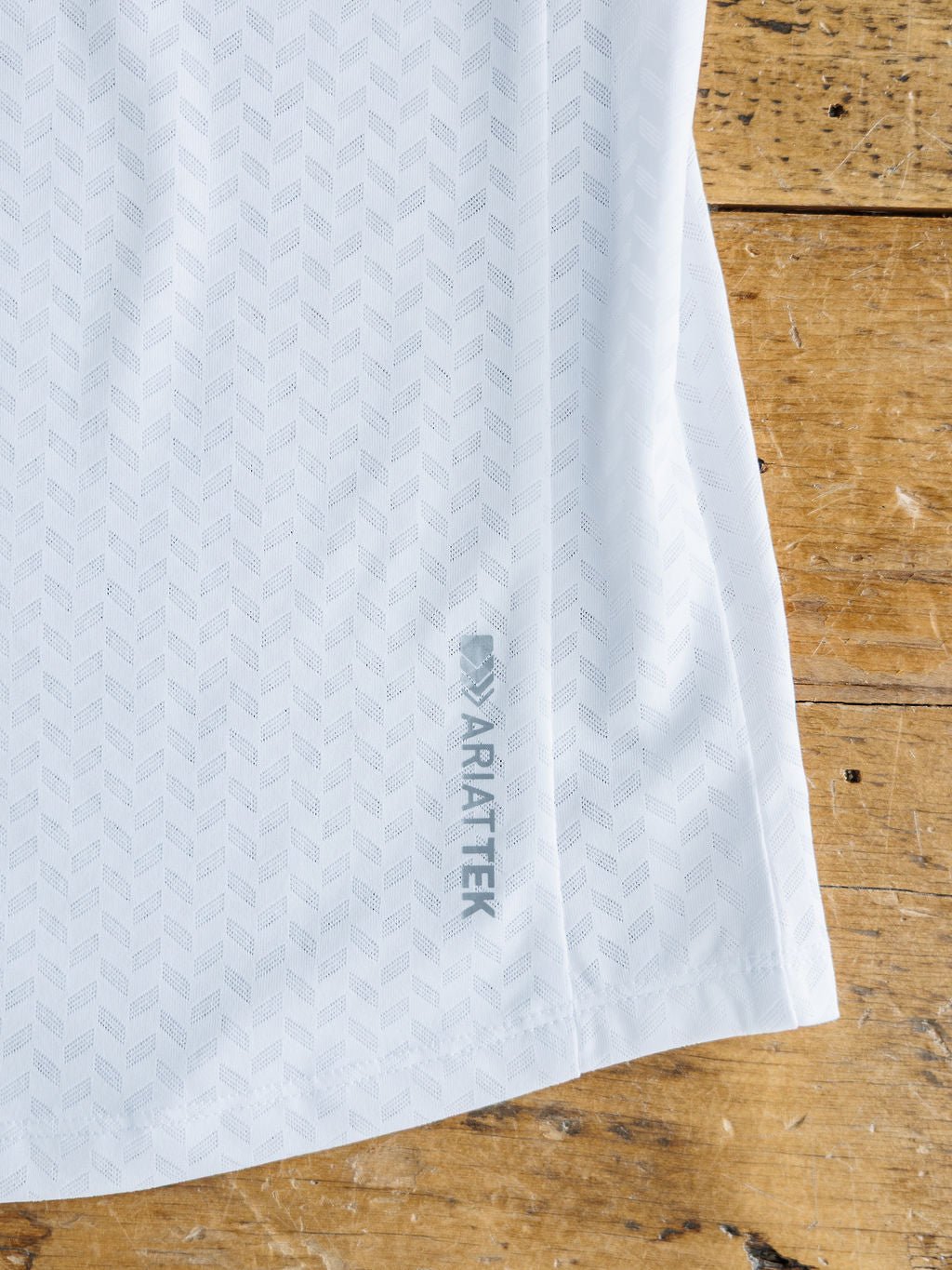 A close-up of the Hanover Ariat White Golf Tank by Savannah Sevens western life{&}style, showing its subtle chevron pattern and ARIAT TEK text near the hem, made from breathable cooling fabric on a weathered wooden surface.