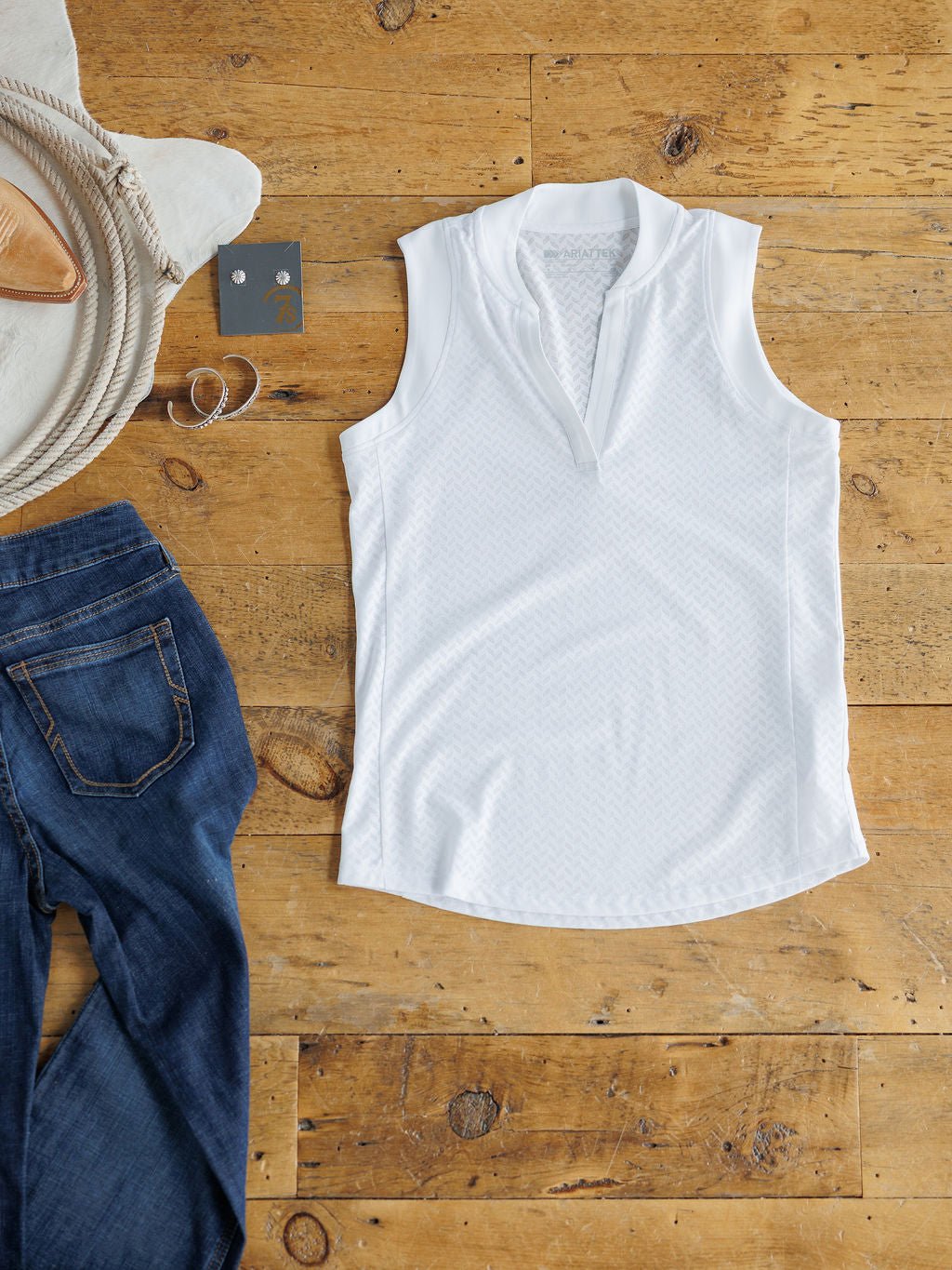 The Hanover Ariat White Golf Tank by Savannah Sevens western life{&}style is displayed flat beside folded blue jeans, a round cream bag, a silver bracelet, and a card with silver earrings on a wooden floor.