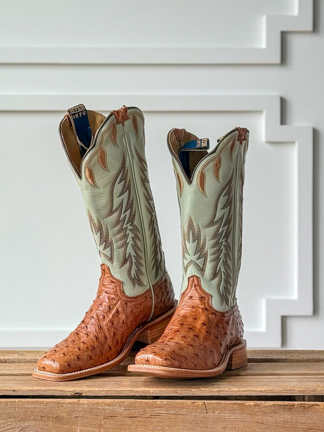 A pair of Harper Ostrich Robin’s Egg Western Leather Boots by Savannah Sevens western life{&}style, featuring a square toe design with brown ostrich leather feet and light green, intricately stitched shafts, displayed on a wooden surface with a white decorative wall in the background. Boots by Hyer.