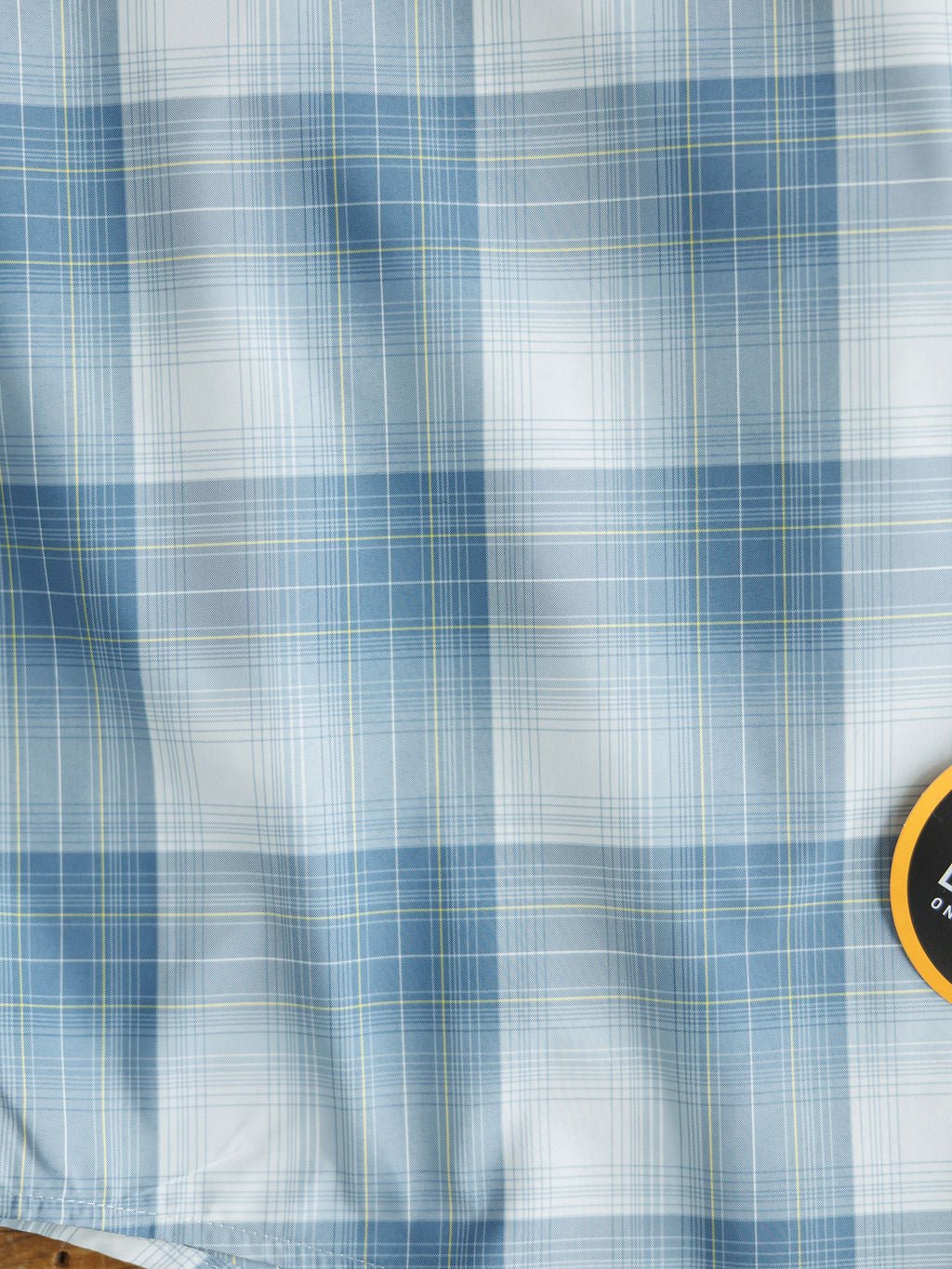 Close-up of blue, white, and light green plaid fabric with varied lines from the Harrison Ariat Men's Blue Plaid Button Up by Savannah Sevens western life{&}style; a small round yellow and black tag is partly visible in the bottom right corner.