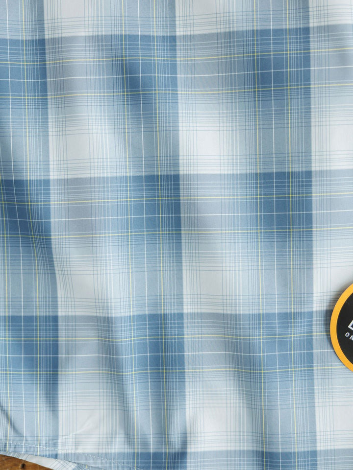 Close-up of blue, white, and light green plaid fabric with varied lines from the Harrison Ariat Men's Blue Plaid Button Up by Savannah Sevens western life{&}style; a small round yellow and black tag is partly visible in the bottom right corner.