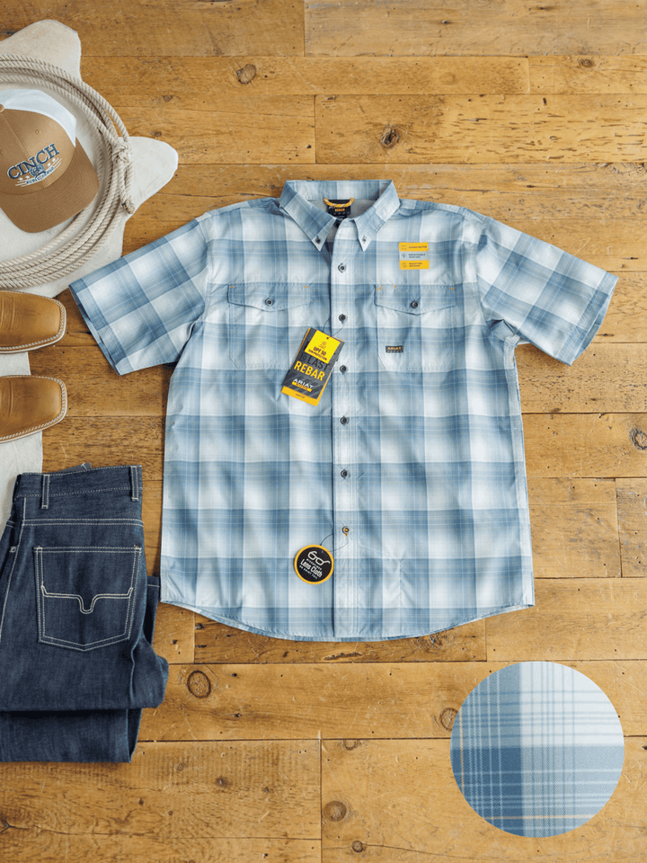 The Harrison Ariat Men's Blue Plaid Button Up from Savannah Sevens western life{&}style is laid flat on a wooden floor with jeans, brown shoes, and a light hat. A closeup showcases the shirt's moisture-wicking blue plaid pattern.