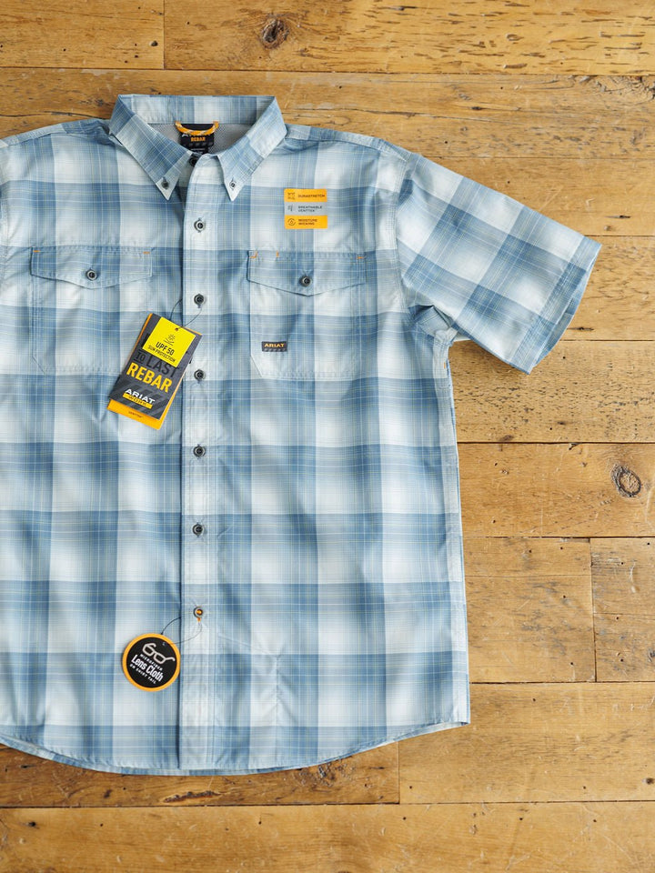 The Harrison Ariat Men's Blue Plaid Button Up by Savannah Sevens western life{&}style is displayed on a wooden surface, featuring two chest pockets, branded labels, and tags attached.