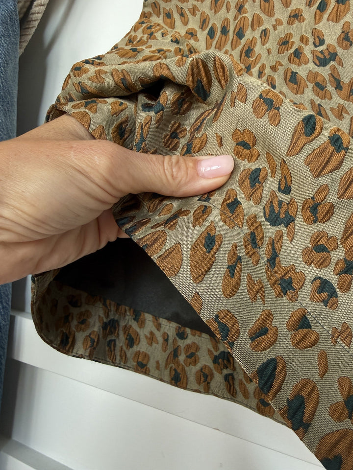 A hand with light pink nail polish gently lifts the edge of the Havana Textured Leopard Sleeveless Top by Savannah Sevens western life{&}style, showcasing its tan fabric with a brown and black leopard pattern. Denim and light knit materials appear blurred in the background.