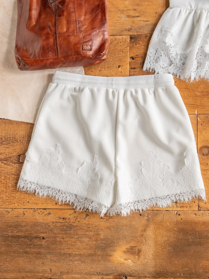 The Havasu Set by Savannah Sevens western life{&}style features white high-waisted shorts with a lace scalloped hem, shown on a wooden surface beside a brown leather bag and matching sleeveless lace top.