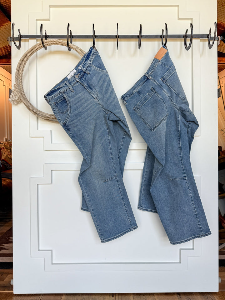 Two pairs of The Hawkin High Rise Barrel Jean by Savannah Sevens western life{&}style, in blue and medium wash denim, hang on black hooks attached to a white paneled door, with wood flooring visible in the background.