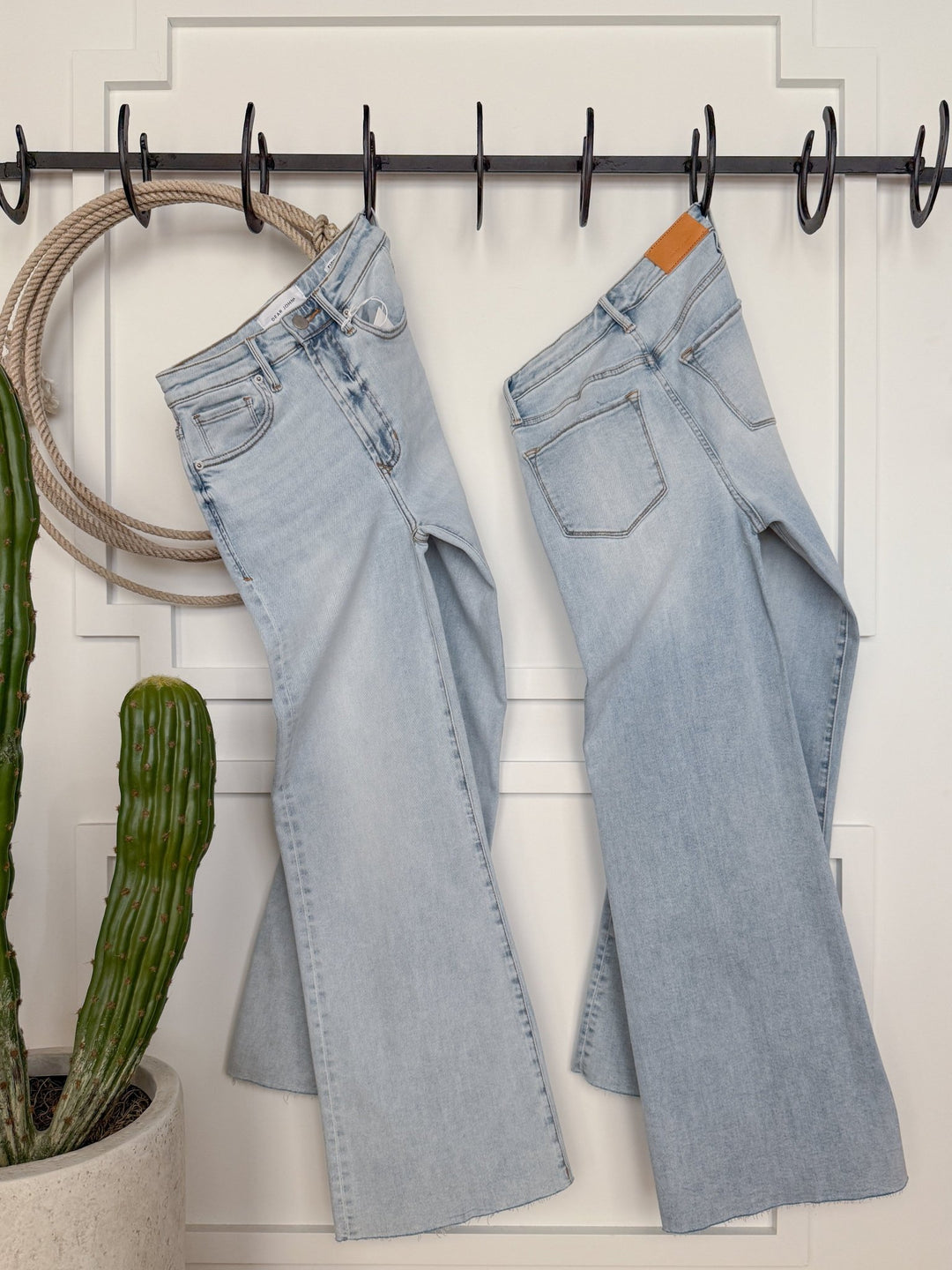 Two pairs of The Hayes High Rise Wide Leg Jeans by Savannah Sevens western life{&}style, in light wash denim, hang side by side from black hooks on a white wall. A potted cactus sits to the left, with a coiled rope nearby.