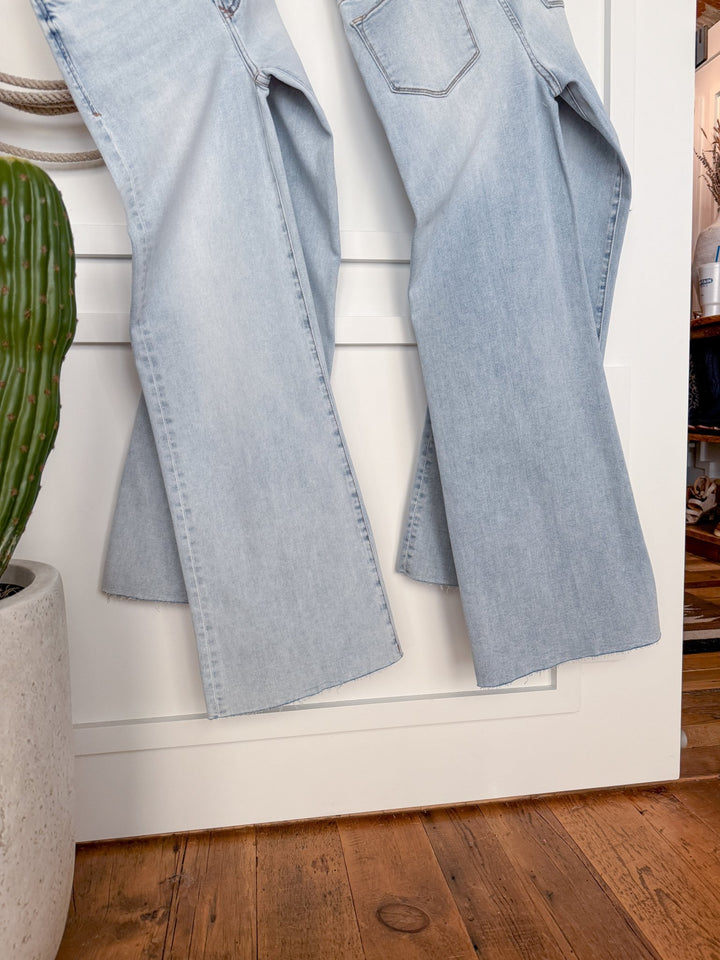 Two pairs of The Hayes High Rise Wide Leg Jean by Savannah Sevens western life{&}style hang side by side against a white paneled wall, with a green potted cactus on the left and wooden flooring visible below.