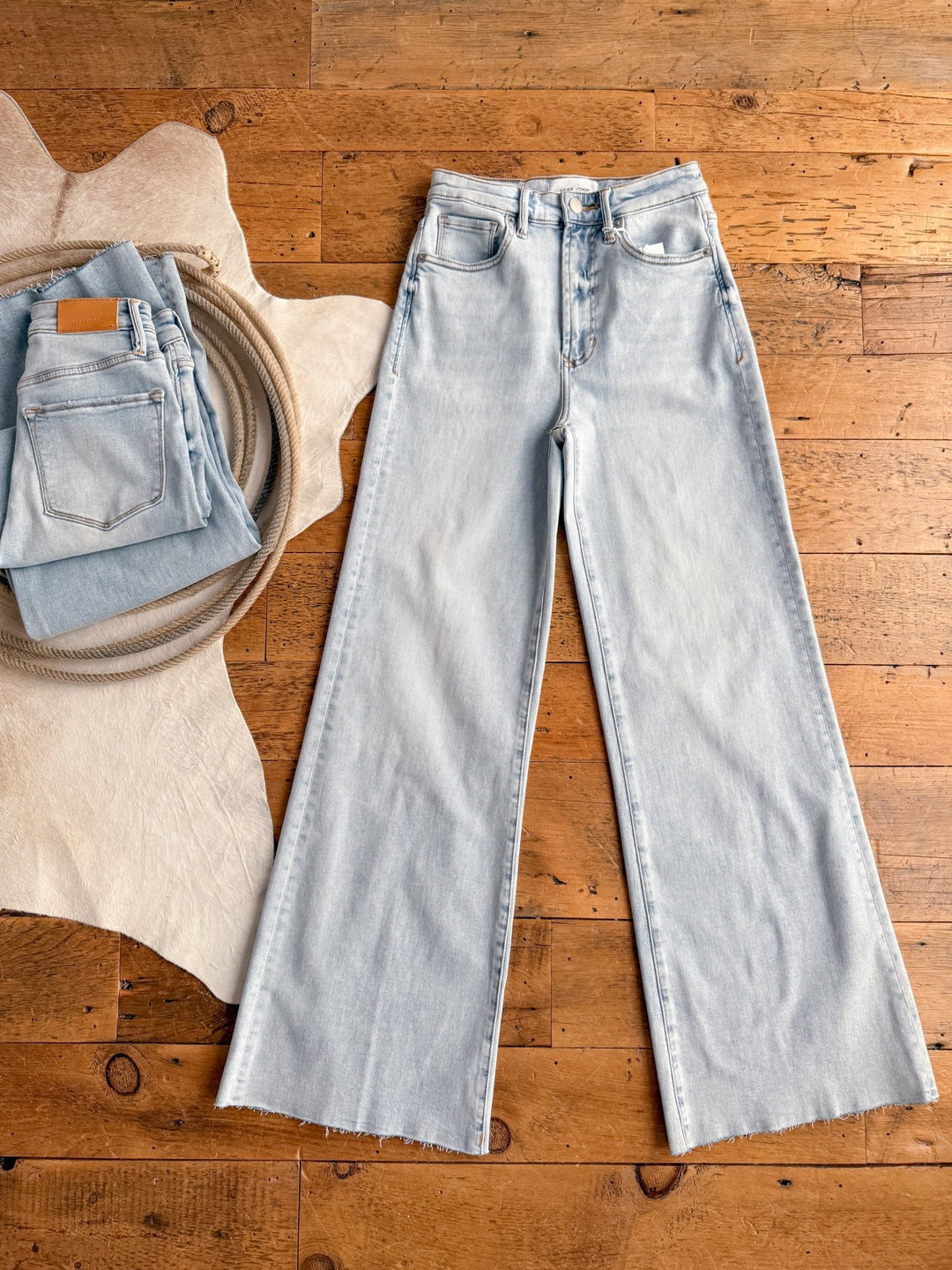 The Hayes High Rise Wide Leg Jean by Savannah Sevens western life{&}style is laid flat on a wooden floor, with a folded pair beside it on a beige textured mat and a cowhide rug.