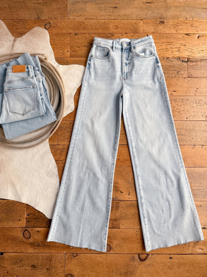 The Hayes High Rise Wide Leg Jean by Savannah Sevens western life{&}style is laid flat on a wooden floor, with a folded pair beside it on a beige textured mat and a cowhide rug.