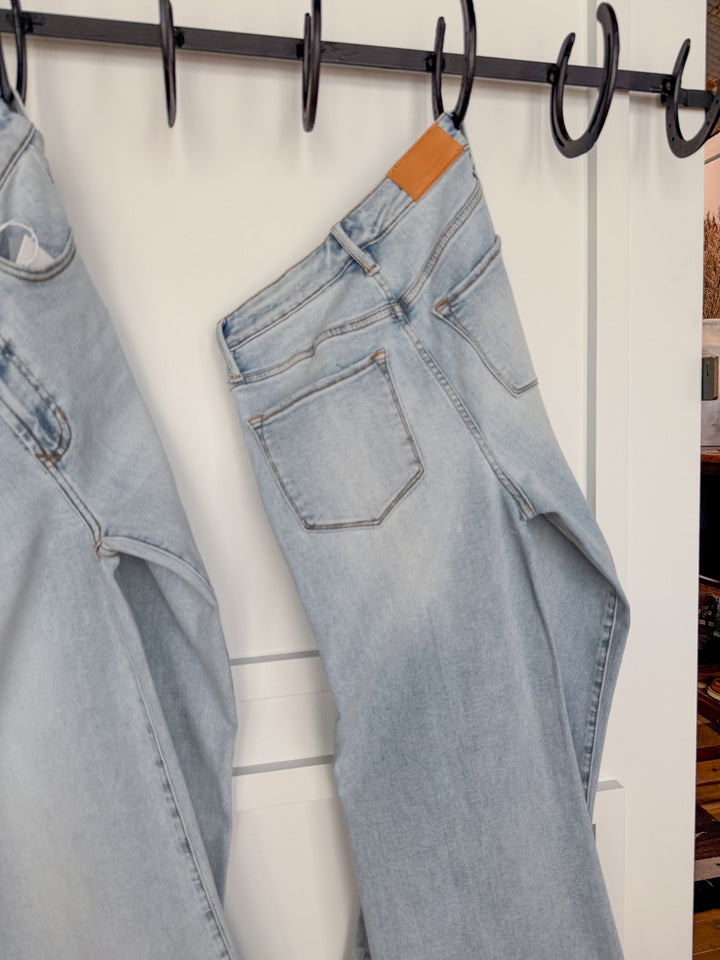 The Hayes High Rise Wide Leg Jean by Savannah Sevens western life{&}style hangs by a belt loop on a black hook against a white wall, with another pair of wide leg jeans partially visible on the left.