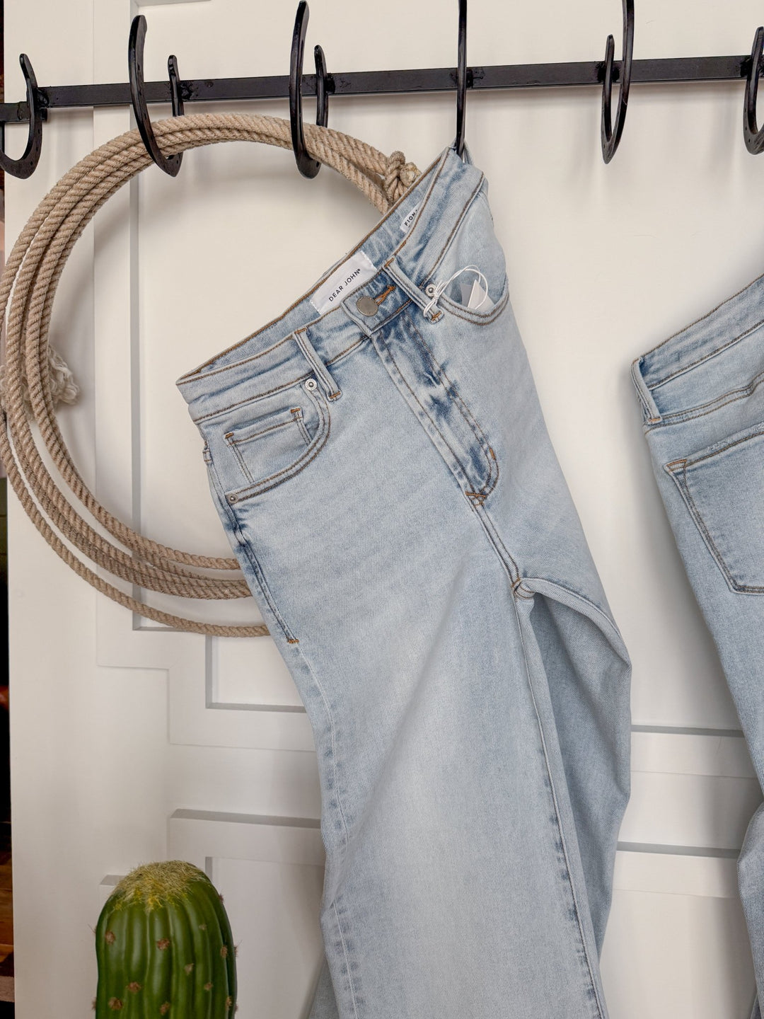 The Hayes High Rise Wide Leg Jean by Savannah Sevens western life{&}style hangs on a black hook, with a coiled rope on the wall behind and a small cactus at the bottom left of the image.