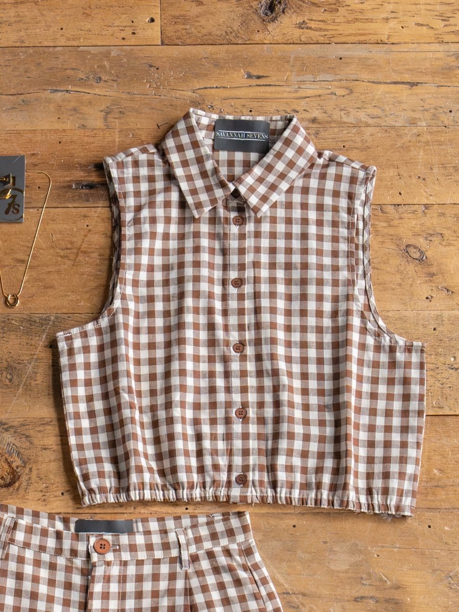 The Hazelton Chocolate Gingham Set by Savannah Sevens western life{&}style features a sleeveless, collared top and elastic waist shorts in brown and white checks, displayed on a wooden surface with the matching shorts peeking from below.