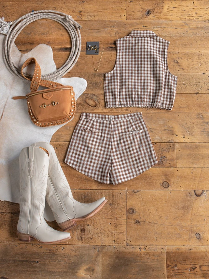 A flat lay featuring the Hazelton Chocolate Gingham Set by Savannah Sevens western life{&}style—sleeveless top and shorts, paired with white cowboy boots, a tan studded crossbody bag, a necklace, and a cowhide rug—ideal for summer.