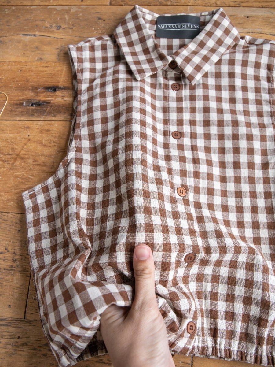 A hand holds the Hazelton Chocolate Gingham Set by Savannah Sevens western life{&}style, showing a hidden pocket in the side seam of this women's cotton top, laid out on a wooden surface.